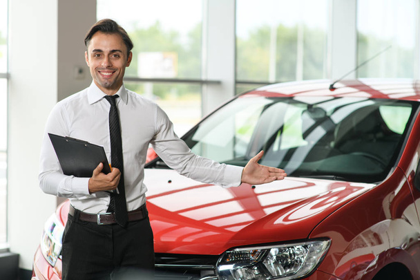 Salesman stay near car. He is waiting for clients. Car is red. He holds tablet in hand. He is smiling. He shows with his hand on brandnew car - Foto, Bild