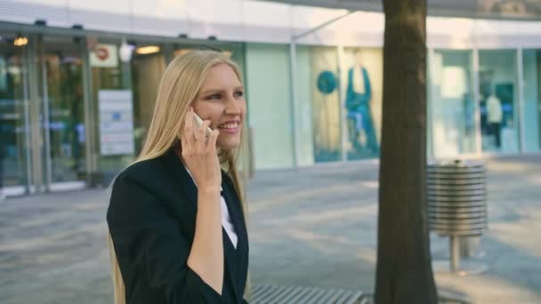 Adult blond businesswoman in suit sitting on bench and waving with hand greeting colleague on street - Кадри, відео
