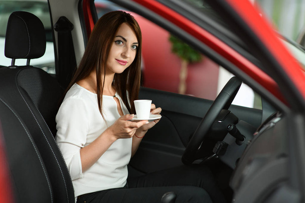 Beautiful girl is drinking coffee in the new car. She is in dealership looking for new car. Car is red. She is smiling. - Zdjęcie, obraz