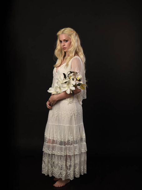 full length portrait of blonde girl wearing white lace dress. standing pose, holding flowers. black studio background. - Foto, afbeelding