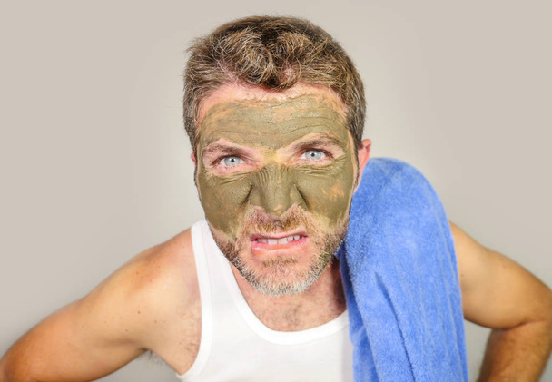 young messy funny man looking at himself horrified in bathroom mirror with green cream on his face applying beauty facial mask product feeling it disgusting and ugly in male cosmetic concept - Fotografie, Obrázek