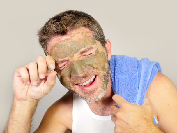lifestyle isolated portrait of young attractive and happy man with bathroom towel  laughing of himself in the mirror with green cream on his face applying facial mask and male skin care cosmetics - Foto, immagini