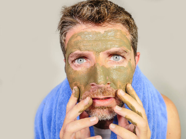 young messy funny man looking at himself horrified in bathroom mirror with green cream on his face applying beauty facial mask product feeling it disgusting and ugly in male cosmetic concept - Foto, Imagen