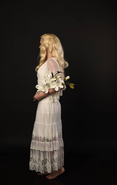 full length portrait of blonde girl wearing white lace dress. standing pose, holding flowers. black studio background. - Fotó, kép