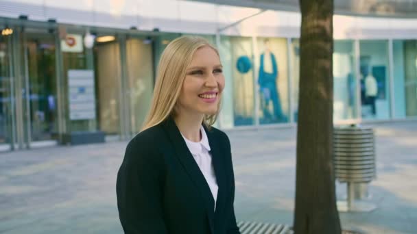 Cheerful executive woman waving with hand. Adult blond business woman in suit sitting on bench and waving with hand greeting colleague on street. - Footage, Video