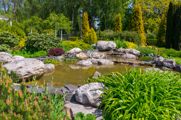 岩、花、緑の茂み、針葉樹と広葉樹に囲まれた装飾的な池 - 写真・画像