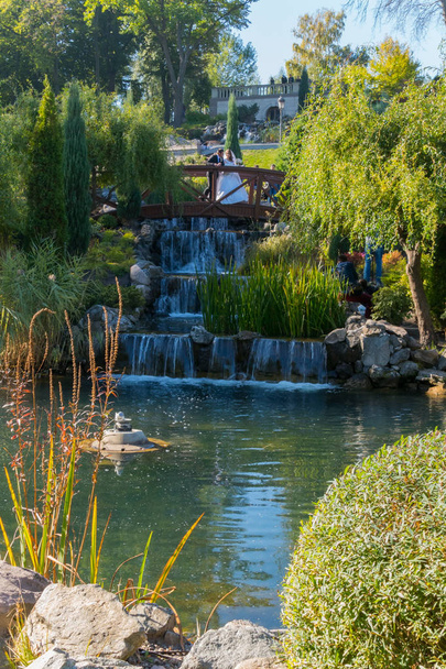 Frischvermählte auf einer Holzbrücke vor dem Hintergrund eines dekorativen Wasserfalls und einer grünen Parkzone - Foto, Bild