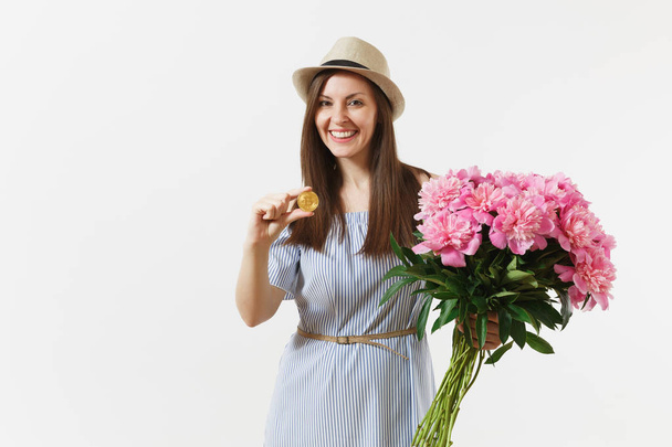 Young woman in dress, hat holding bitcoin, coin of golden color, bouquet of beautiful pink peonies flowers isolated on white background. Business, delivery, online shopping, virtual currency concept - Foto, imagen