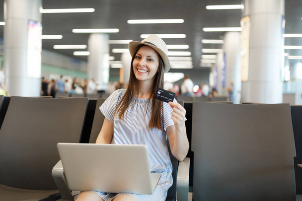 Молодая улыбчивая туристка в шляпе работает на ноутбуке, держа кредитную карточку, ожидая в холле международного аэропорта. Пассажир, путешествующий за границу по выходным. Воздушные перевозки, концепция полета
. - Фото, изображение