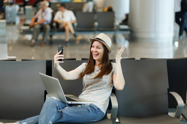 Rire voyageur touriste femme travaillant sur ordinateur portable, faire selfie sur téléphone mobile, écarter les mains, attendre dans le hall d'entrée à l'aéroport. Passager voyageant à l'étranger le week-end escapade. Concept de vol aérien
 - Photo, image