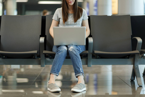 Die junge lächelnde Touristin arbeitet am Laptop, während sie in der Lobby des internationalen Flughafens wartet. Passagiere, die am Wochenende im Ausland unterwegs sind. Flugreisen, Flugreise-Konzept - Foto, Bild