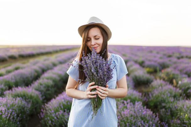 Young sensual beautiful woman in blue dress, hat on purple lavender flower blossom meadow field outdoors on summer nature background. Tender female near flowering bush with bouquet. Lifestyle concept - Foto, afbeelding