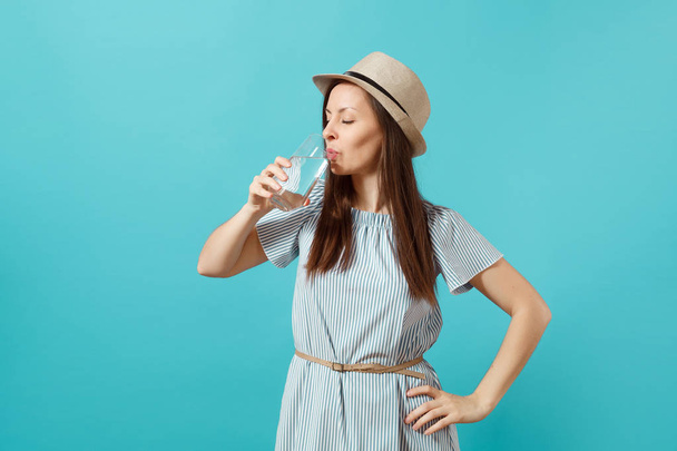 Portrait of young beautiful woman in blue dress, hat holding and drinking clear fresh pure water from glass isolated on blue background. Healthy lifestyle, people sincere emotions concept. Copy space - Foto, Bild
