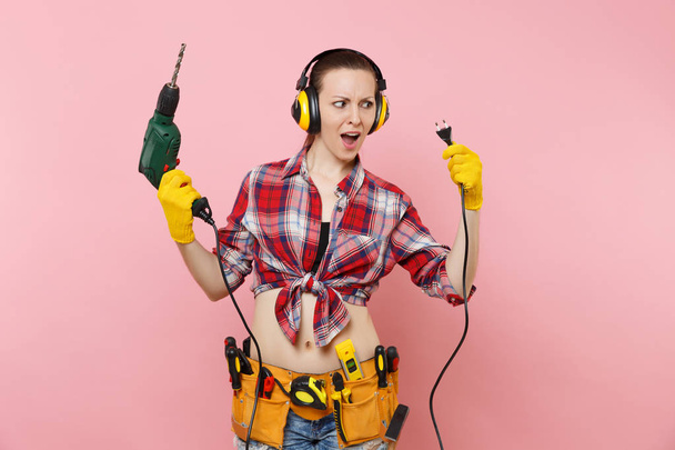 Ενέργεια πολυτεχνίτης σέξι γυναίκα στα γάντια, θόρυβος μονωμένη ακουστικά, κιτ εργαλείων ζώνη γεμάτη μέσα κρατώντας ηλεκτρικό δράπανο, απομονώνονται σε ροζ φόντο. Θηλυκό σε αρσενικό το έργο. Ανακαίνισης - Φωτογραφία, εικόνα