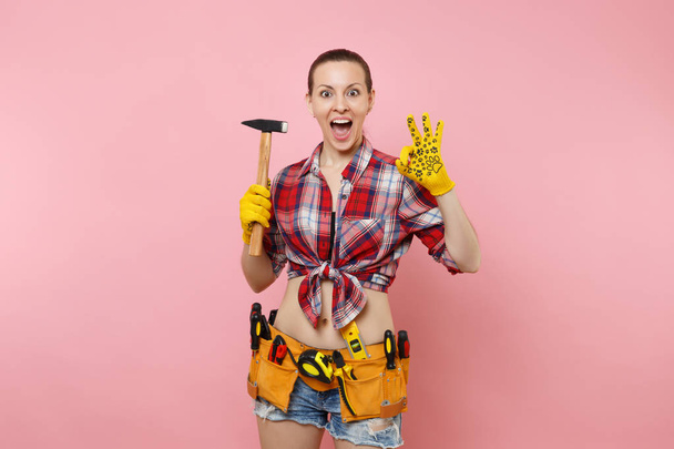 Strong young handyman woman wearing plaid shirt, denim shorts, kit tools belt full of different instruments hammer isolated on pink background. Female in male work. Renovation and occupation concept - Foto, imagen