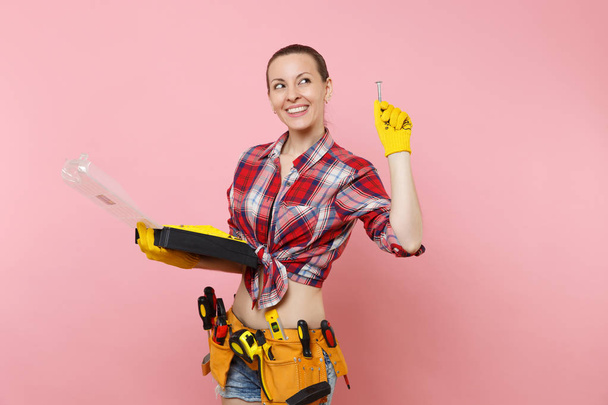 Strong young handyman woman in plaid shirt, denim shorts, kit tools belt full of different instruments holding toolbox isolated on pink background. Female in male work. Renovation occupation concept - Photo, Image