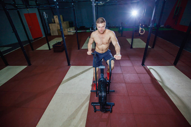 Fit young man using exercise bike at the gym. Fitness male using air bike for cardio workout at cross training gym. - Valokuva, kuva