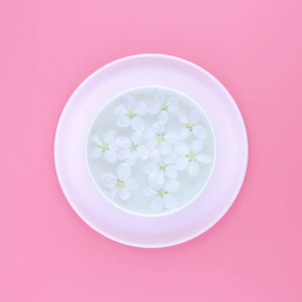 Concept créatif plat vue du dessus du bol d'arôme avec de l'eau et des fleurs de pommier fleurissant au printemps blanc sur fond carré en papier rose pastel avec espace de copie dans un style minimal, modèle pour le texte
 - Photo, image