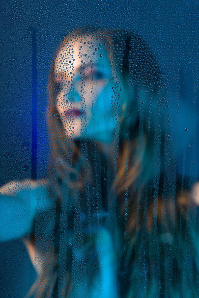 Art style portrait in blue tones of a lonely beautiful sad model girl behind a window glass, over which rain drops down. Artistic blur on model face. Conceptual, commercial and fashionable. Copy space - Foto, afbeelding