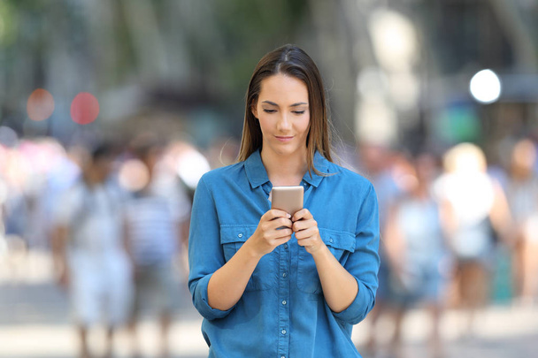 Front view of a serious woman in blue using a smart phone walking on the street - Photo, Image