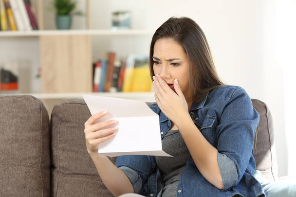 Грустная женщина читает плохие новости в бумажном письме, сидя на диване в гостиной дома
 - Фото, изображение