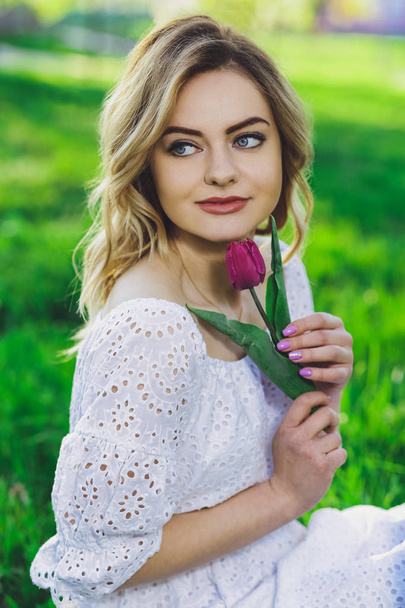Young beautiful blonde girl in white dress with a flower tulip on a background of green grass - Foto, Imagem
