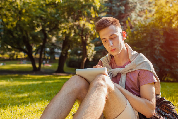 Handsome college man writing notes in copybook in campus park. Happy guy student learning outdoors sitting on grass in spring. Education - Zdjęcie, obraz