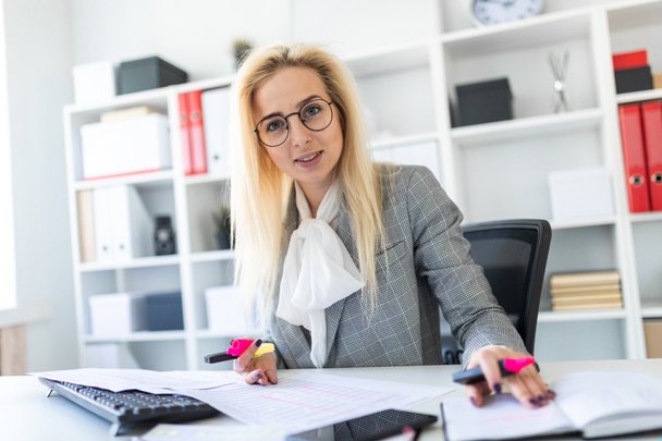 Μια νεαρή κοπέλα με τα γυαλιά που εργάζεται στο γραφείο με ένα δείκτη, ένα σημειωματάριο και έγγραφα. - Φωτογραφία, εικόνα