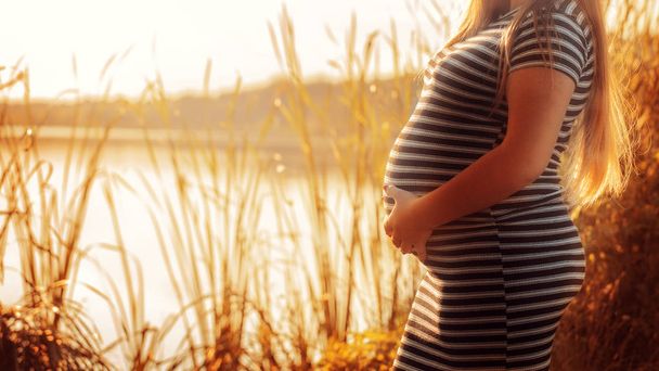 Hamile kadının karnı sunset.close-up bir nehir arka plan üzerinde çizgili bir elbise içinde hamile bir kadın, çok dokunaklı - Fotoğraf, Görsel