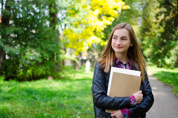 Portrait of smiling university student standing in the park on her way to class. Girl is wearing a backpack and holding textbooks. Young adult high school or college student. Back to school concept. - Foto, Bild