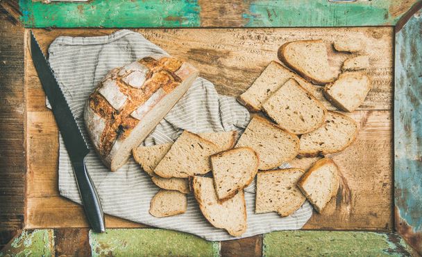 Freshly baked sourdough wheat bread loaf halved and cut in slices on board over linen napkin and rustic wooden tray background - Foto, Imagen