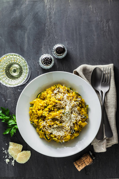 Italian dish yellow Risotto milanese with saffron, zucchini and Parmesan cheese on a black slate table with white wine in a glass. north Italy kitchen - Fotó, kép