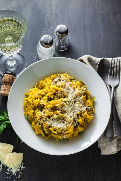 Italian dish yellow Risotto milanese with saffron, zucchini and Parmesan cheese on a black slate table with white wine in a glass. north Italy kitchen - Valokuva, kuva