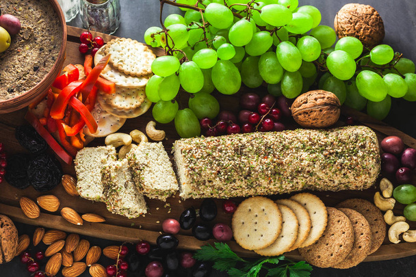 Wooden serving board with vegan snacks. Pate of olives and cheese from cashew nuts with herbs. Healthy appetizer with grapes, nuts, and dried fruits on a black slate table. - Photo, Image