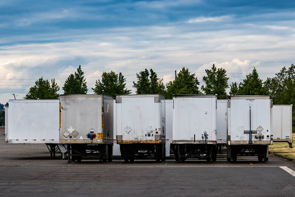 Semi tractor trailers parked in a row with doors closed under a blue cloudy sky with trees - Photo, Image