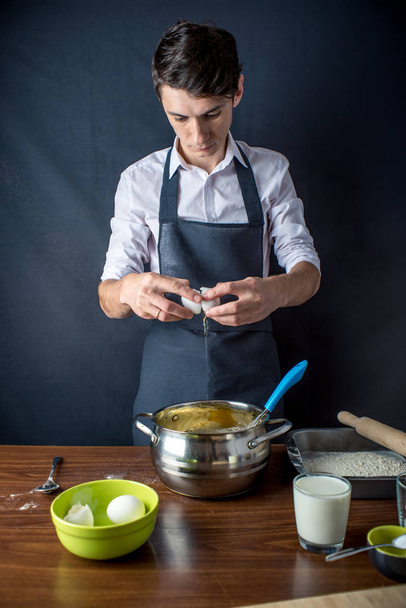 Mladý muž, kuchař v jednotné černé zástěře na vaření dortu s přísadami na stole v kuchyni. Koncept muži vaří dezerty - Fotografie, Obrázek