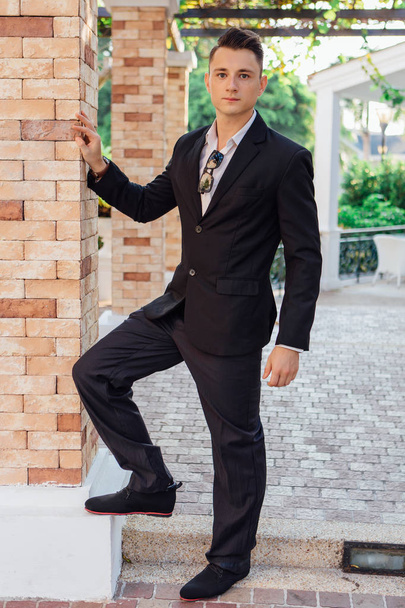 Young stylish handsome man posing in modern city park, wearing suit and sunglasses. - 写真・画像