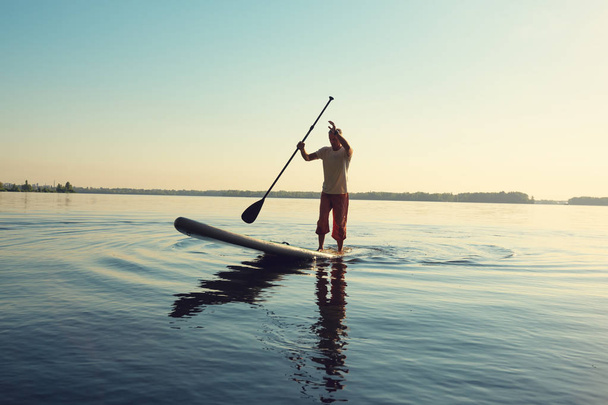 Man is training on a SUP board on a large river during sunny morning. Stand up paddle boarding - awesome active recreation in nature. - Photo, Image