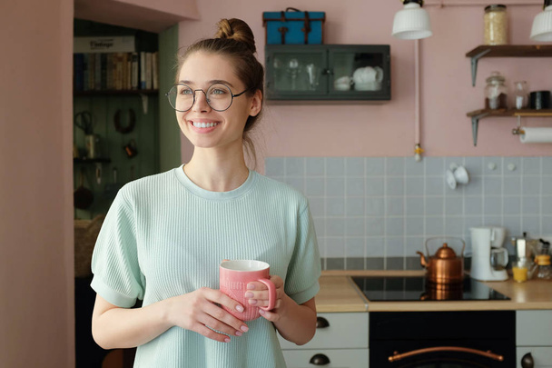 Imagem de perto de jovens otimista europeu caucasiano feminino passar a tarde em sua cozinha segurando xícara de bebida nas mãos com olhar sonhador, antecipando bom dia frutífero no trabalho ou em casa
 - Foto, Imagem