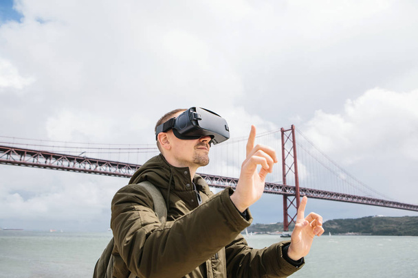 Ένας άνθρωπος χρησιμοποιεί γυαλιά εικονικής πραγματικότητας. 25η Απριλίου γέφυρα στη Λισαβόνα στο παρασκήνιο. Η έννοια του εικονικού ταξιδιού. Η έννοια των σύγχρονων τεχνολογιών και τη χρήση τους στην καθημερινή ζωή - Φωτογραφία, εικόνα