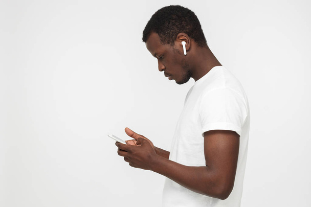 Side shot of young African man isolated on gray background holding smartphone, browsing webpages or looking for new tracks to listen through his new white wireless earphones with concerned face - Foto, afbeelding