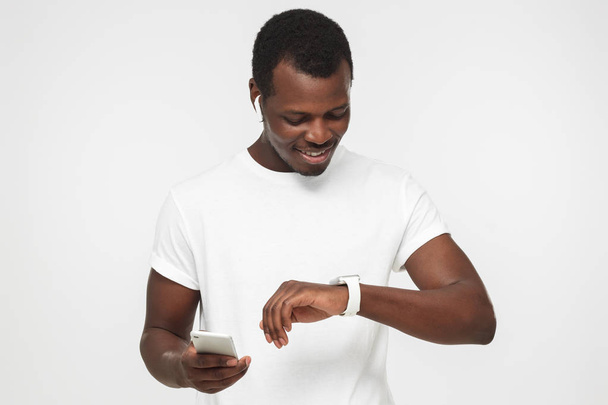 Closeup shot of African American man pictured isolated on grey background listening to music through wireless earphones, using smartphone and looking at digital smart wristwatch, fan of gadgets - Foto, Imagen