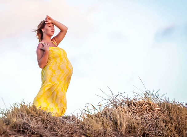Young and attractive Caucasian pregnant woman in a long yellow dress enjoying sunset somewhere outdoors in the nature. Expecting a child, expecting a baby concept, looking forward for child birth idea. - Foto, afbeelding