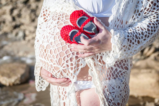 Young pregnant Caucasian woman holding tiny baby girl shoes in her hands and holding her belly. Expecting a girl daughter concept image. Expecting mother, future parent. Pregnant woman belly. - Photo, Image