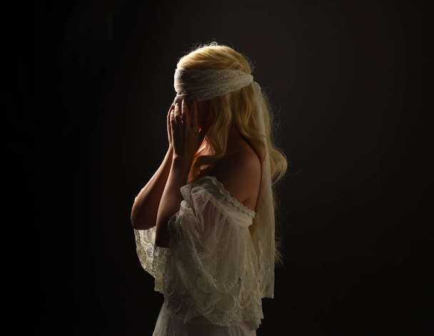 Porträt eines blonden Mädchens in einem weißen Spitzenkleid mit Augenbinde. schwarzer Studiohintergrund. - Foto, Bild