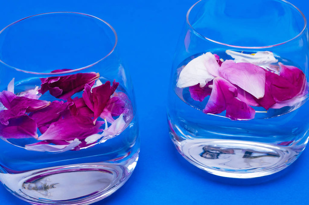 Üveg víz és a pünkösdi rózsa virág szirmai a kék háttér. - Fotó, kép