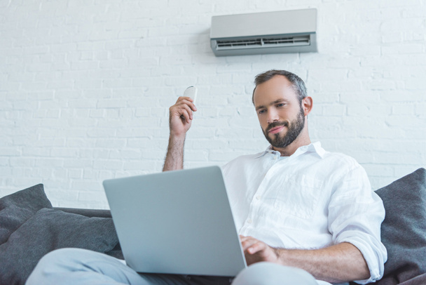 bearded man turning on air conditioner with remote control while using laptop - Photo, Image