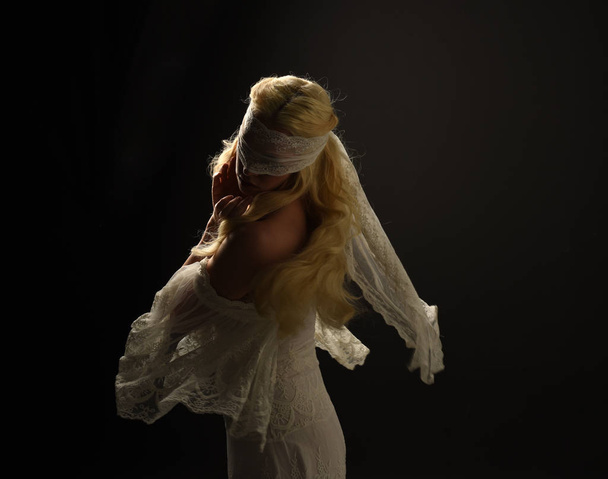 портрет блондинки в белом кружевном платье и с завязанными глазами. черный фон студии
. - Фото, изображение