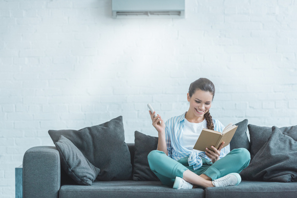 šťastná žena čtení knihy při zapnutí klimatizace s dálkovým ovládáním doma - Fotografie, Obrázek