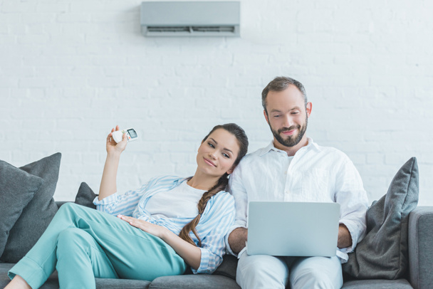 Ehepaar schaltet bei Sommerhitze Klimaanlage ein, während es Laptop benutzt - Foto, Bild